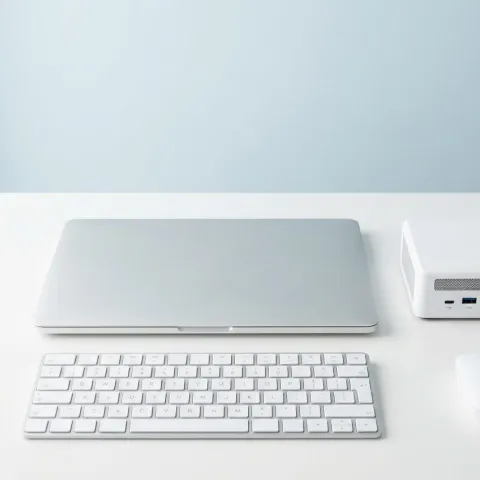 Portátil cerrado, teclado, ratón y miniordenador sobre un escritorio blanco.